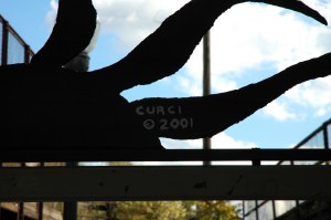 The back of a steel sun shaped sculpture with the artists name and 2001 painted in white on it. Artist name is curci. 
