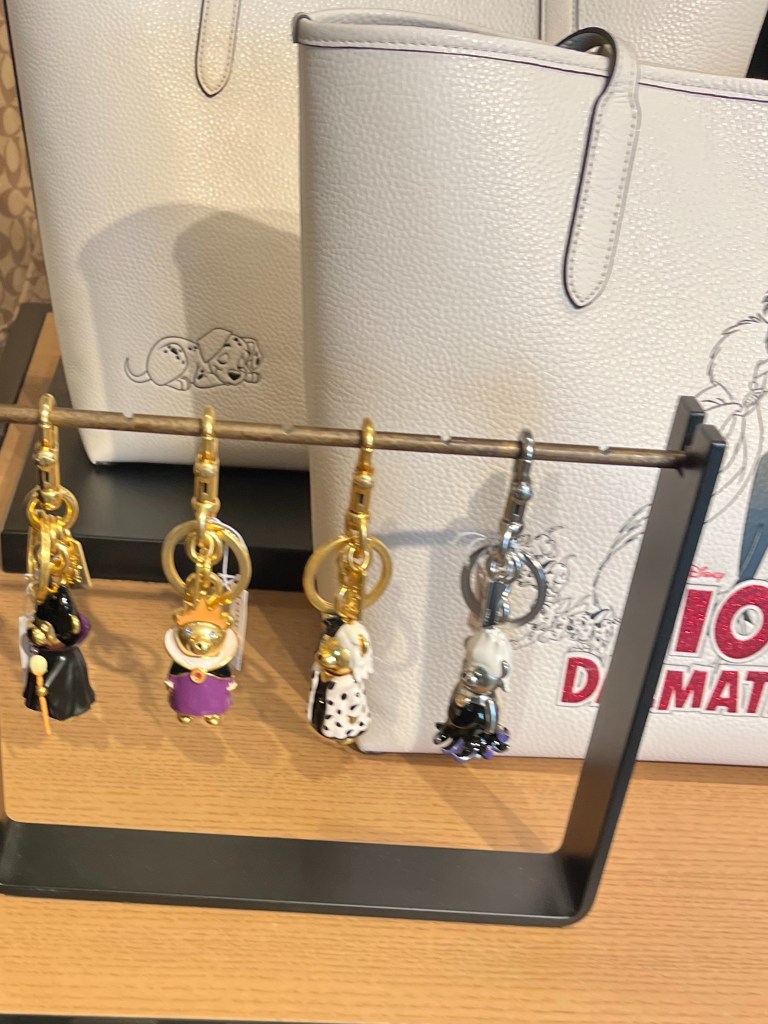 Four gold bear-shaped charms hang on a small tabletop rack. They are dressed like the Old Woman from Snow White, Evil Queen, Cruella, and Ursula.