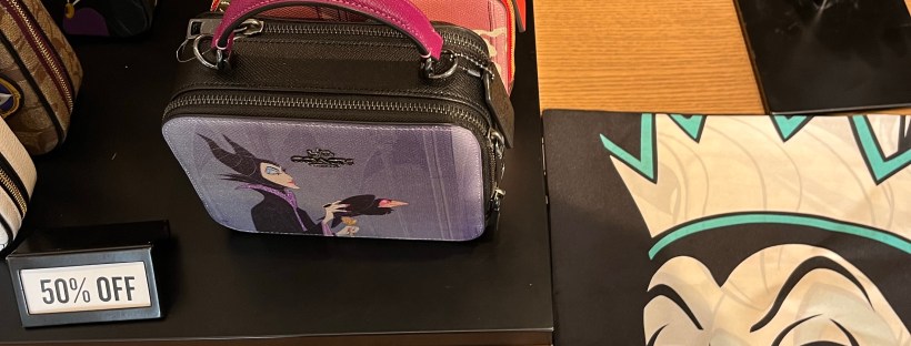 Three Coach designer bags on a table with Disney villian designs on them. Maleficent and the Evil Queen are on two of the bags, and a scarf with the Evil Queen is laid out next to them.